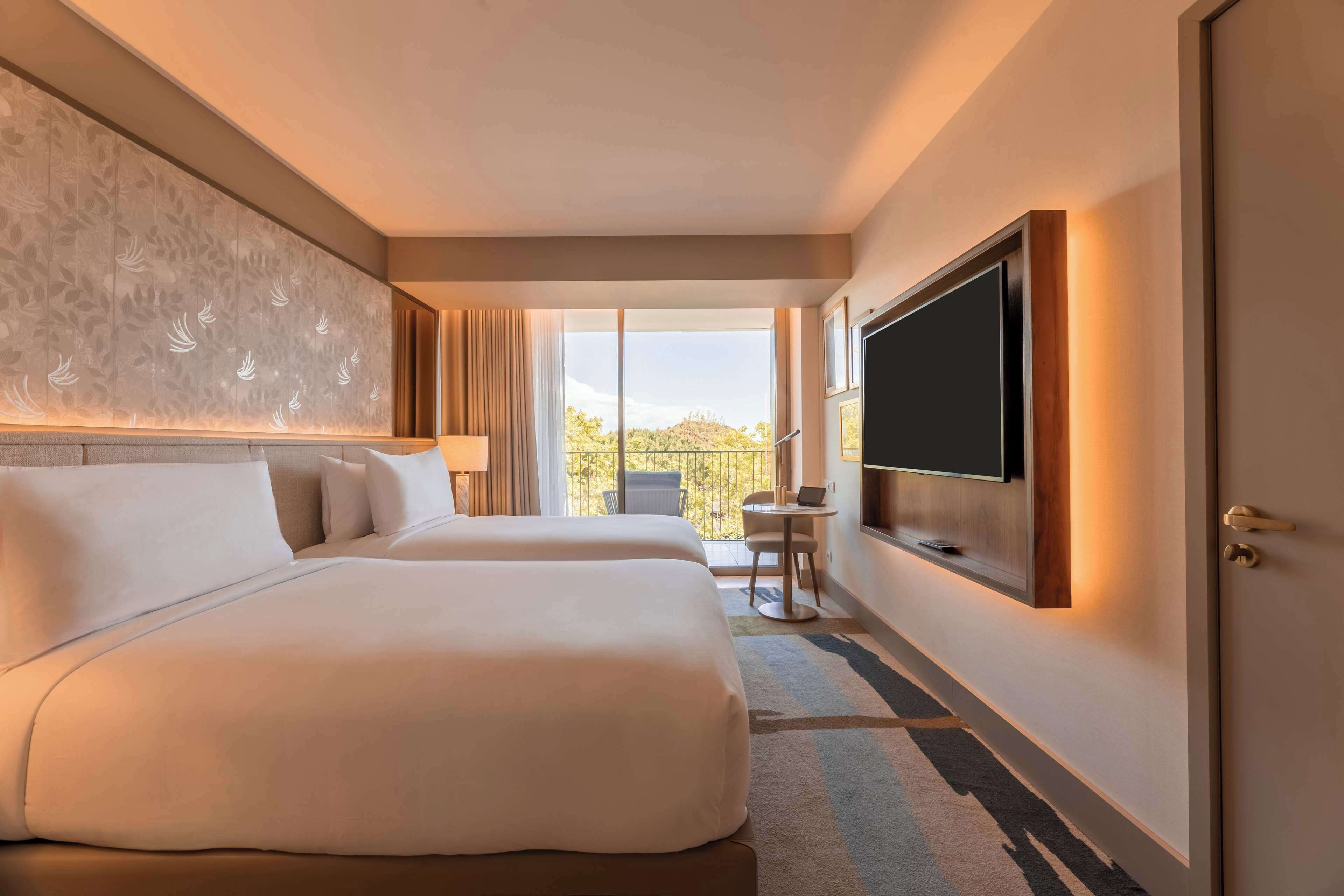 Ein Hotelzimmer mit zwei Betten, einem wandmontierten Flachbildfernseher, einem Balkon mit Blick und einem kleinen Tisch mit Stuhl.