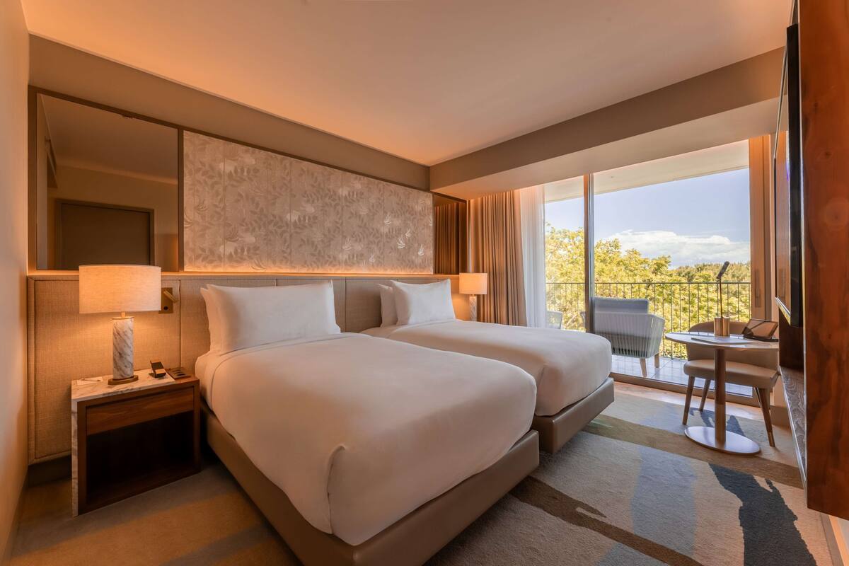 Habitación de hotel con dos camas, un escritorio y un balcón con vistas.