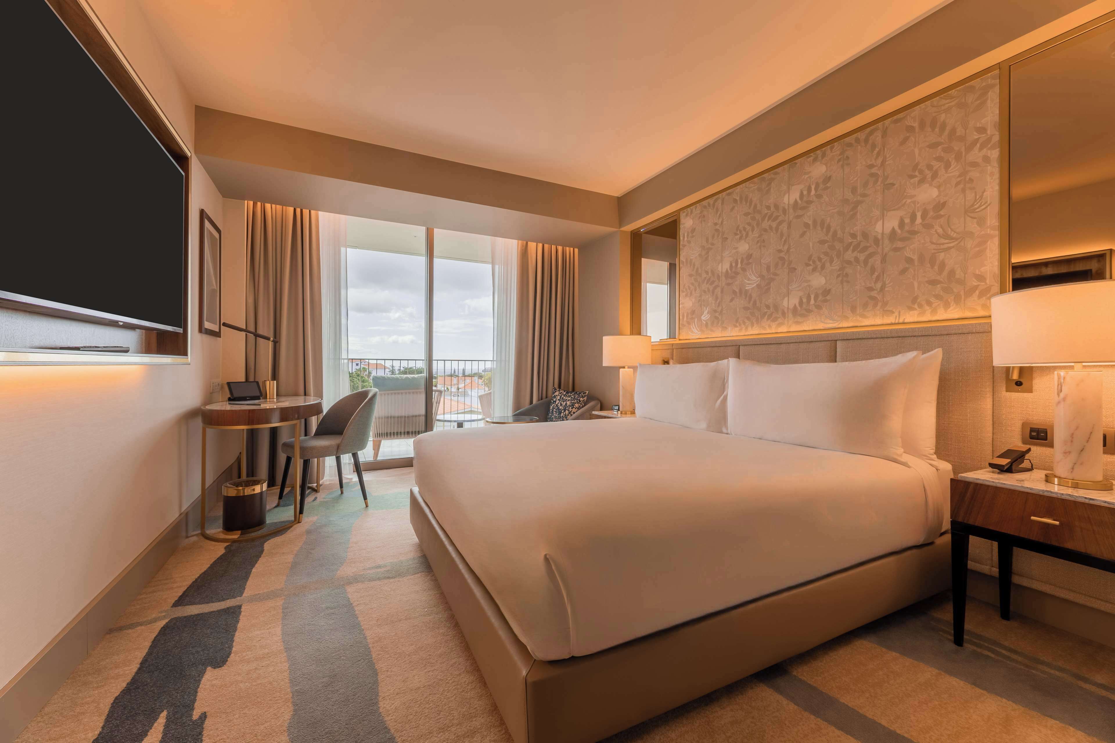 A hotel room with a large bed, a desk with a chair, a TV, and a view of the city through large windows.