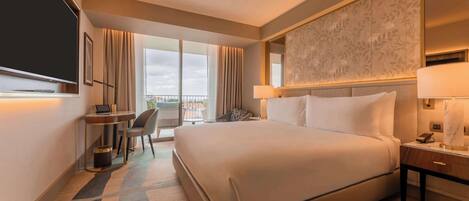 A hotel room with a large bed, a desk with a chair, a TV, and a view of the city through large windows.