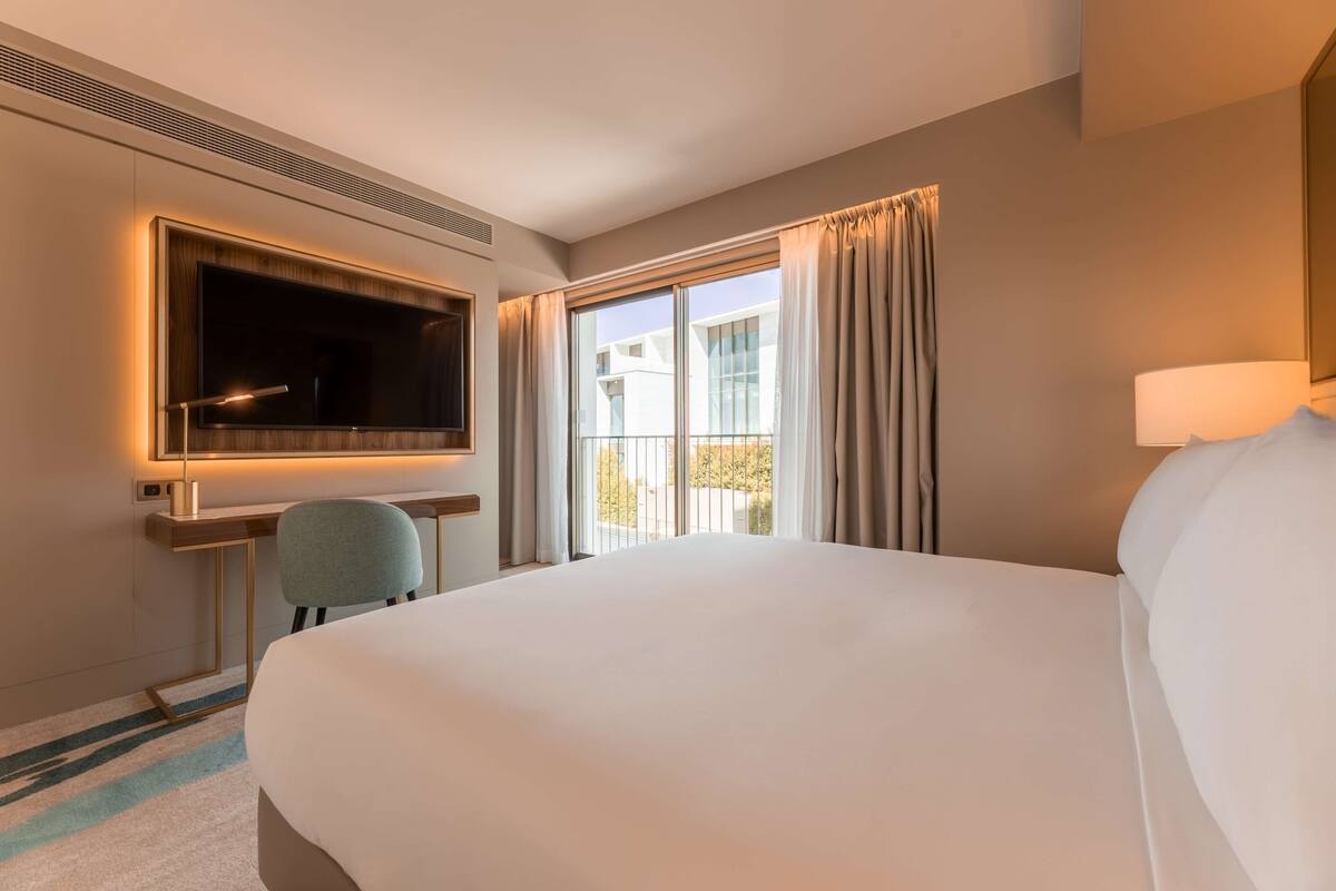 Habitación de hotel con una cama grande, un escritorio con silla, televisor y una ventana con cortinas.