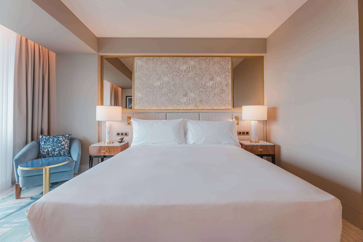 Una habitación de hotel con una cama grande, dos mesitas de noche con lámparas, una silla y un espejo grande sobre la cama.