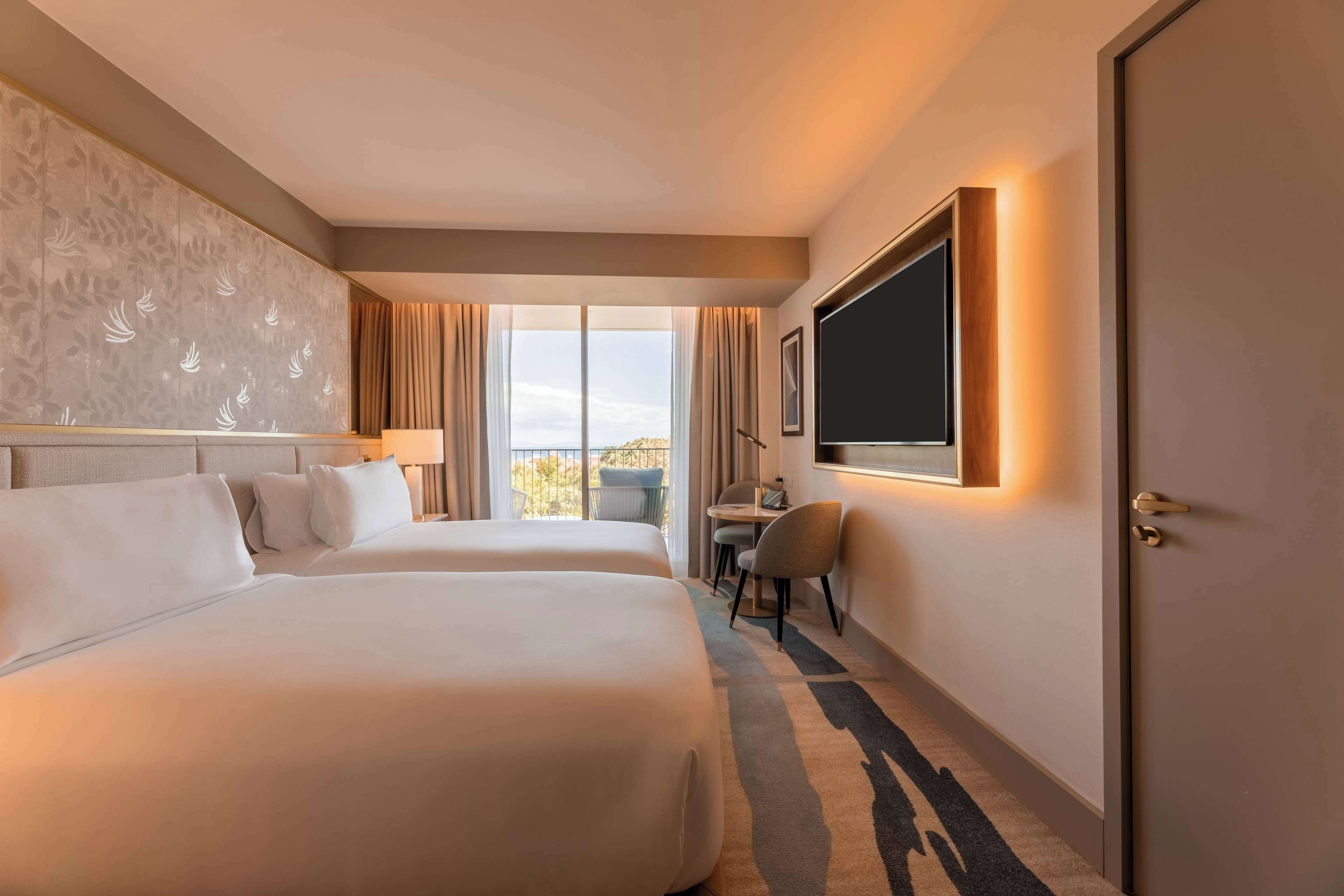 Ein Hotelzimmer mit zwei Betten, einem Schreibtisch, einem Sessel, einem Fernseher und einem großen Fenster mit Vorhängen.