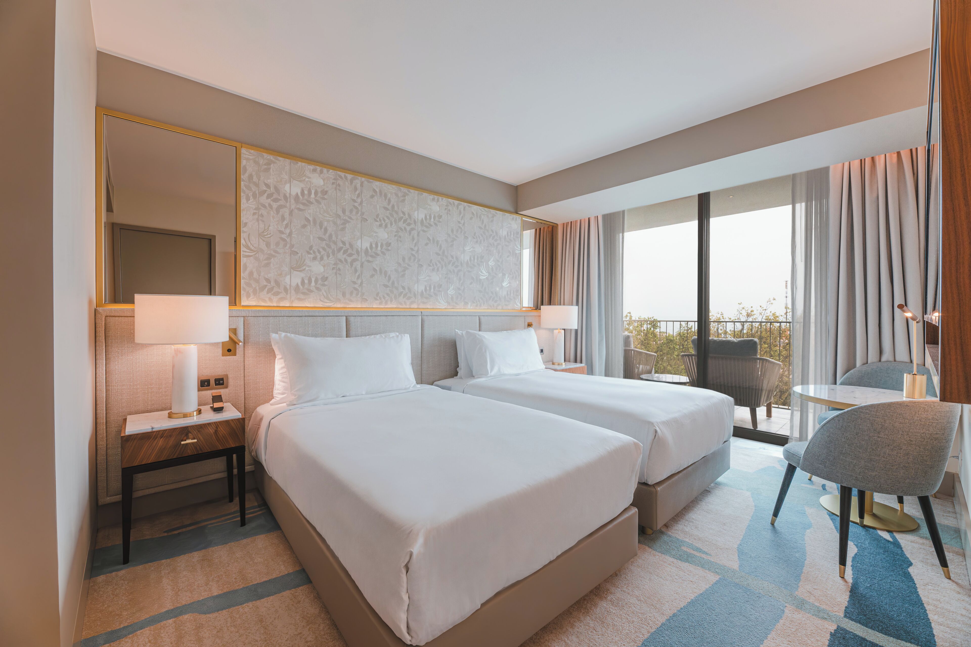 A hotel room with two beds, a sitting area with a table and chair, a balcony with a view, and a large mirror.