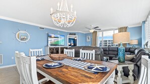 A dining area with a wooden table, white chairs, a chandelier, and a television displaying a scenic view. - Destin West Heron 509 ~ Beach Paradise ~ Overlooking the Lazy River (Fort Walton Beach)