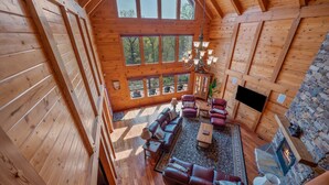 A spacious log cabin interior with a large living area, a stone fireplace, and large windows offering a view of the surrounding forest. - Chateau Laurel in Lake Lure | Private 5,000 Sq Ft Cabin in Blue Ridge Mountains! (Lake Lure)