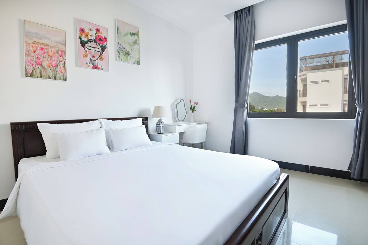 Ein Hotelzimmer mit einem großen Bett, einem Nachttisch mit Lampe, einem kleinen Schreibtisch und einem Fenster mit Blick auf ein Gebäude und Berge.