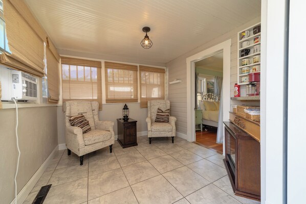 A cozy room with two armchairs, a small table, and a lamp. There are large windows with blinds, a ceiling light, and a shelf with various items.