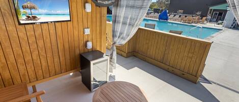 A wooden deck with a pool, lounge chairs, and a TV screen displaying a beach scene.
