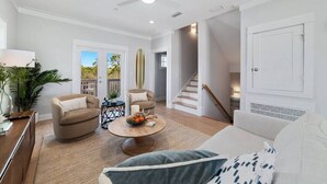 A spacious living room with a ceiling fan, a round coffee table with fruits, a sofa, and a staircase leading to another level. - Summer Vacation! Beach, Pool, Games, Sleeps 10! (Orange Beach)