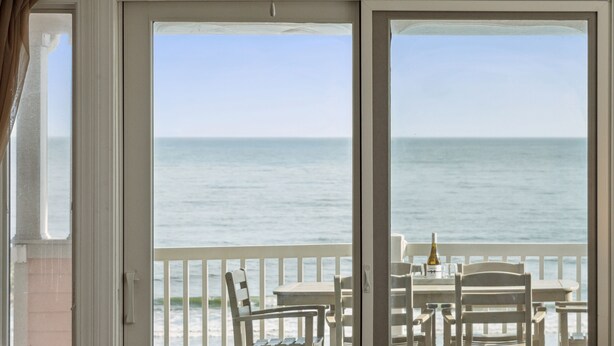 Ein Zimmer mit einer großen Schiebeglaswand, die auf einen Balkon mit Meerblick führt. Auf dem Balkon stehen ein Tisch, Stühle und eine Weinflasche.