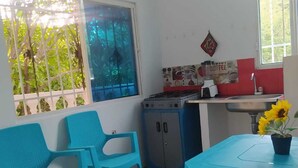 A small kitchen with a gas stove, a sink, and a window with a view of greenery. There are two blue chairs and a table with a sunflower. - APARTMENT WITH BALCONY ECONOMIC, QUIET, IN COUNTRY HOUSE NEAR THE SEA (SANTA MARTA)