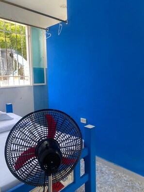 A room with a bed, a standing fan, a window with metal bars, and a blue wall.