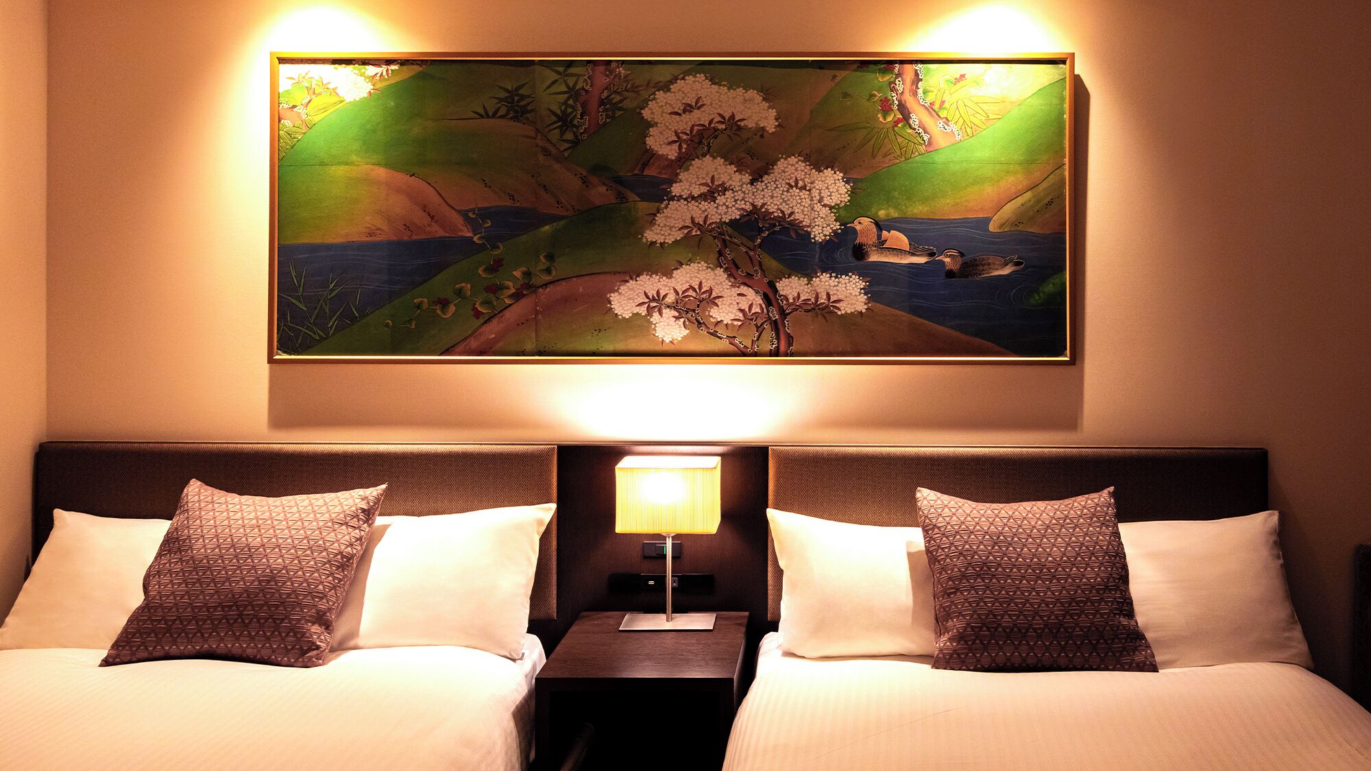 Quarto de hotel com cama, duas almofadas, mesa de cabeceira, abajur e pintura de flores de cerejeira.