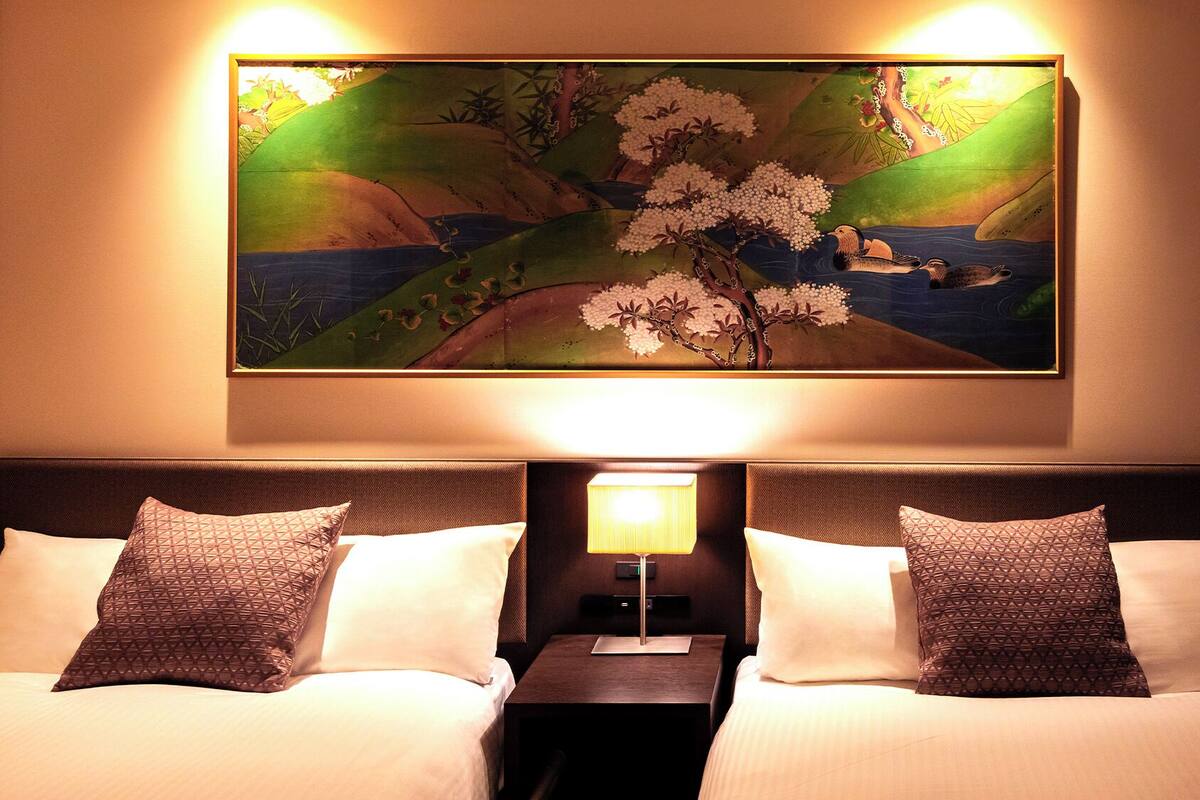 A hotel room with a bed, two pillows, a nightstand, a lamp, and a painting of cherry blossoms.