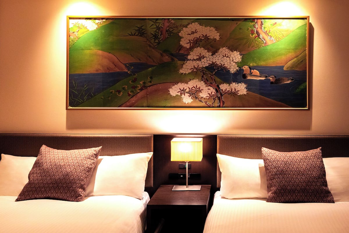 Quarto de hotel com cama, duas almofadas, mesa de cabeceira, abajur e pintura de flores de cerejeira.