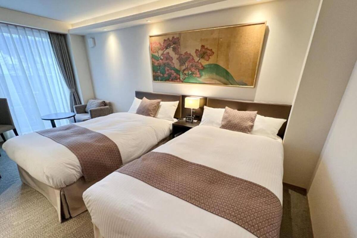 A hotel room with two beds, a painting on the wall, a small table, and a chair.