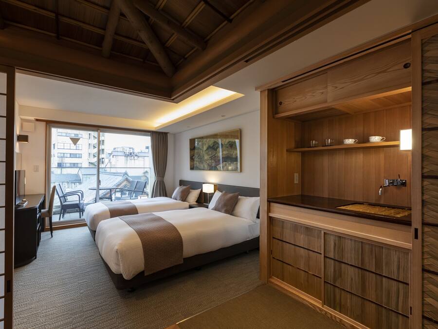 Ein Hotelzimmer mit zwei Betten, einem Schreibtisch, einem Sessel, einem großen Fenster und einem Holzkorpus mit Waschbecken.