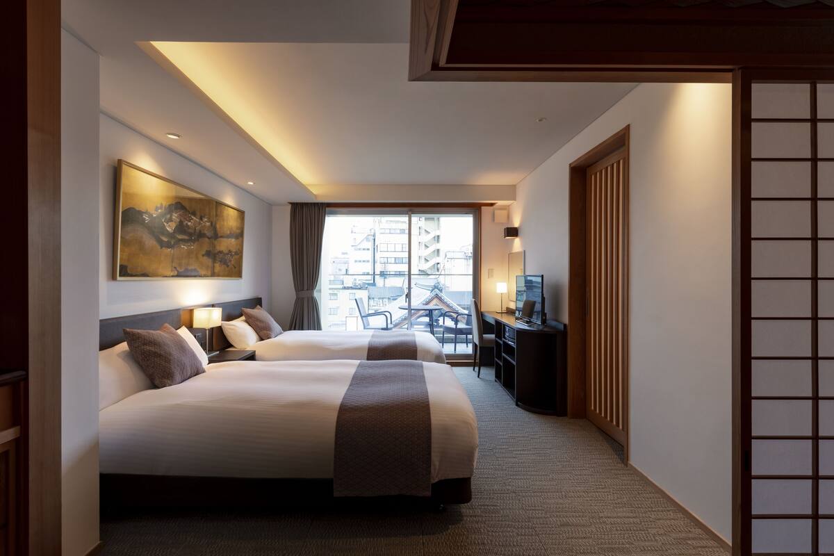 A hotel room with two beds, a desk, a chair, and a large window with a view of buildings.