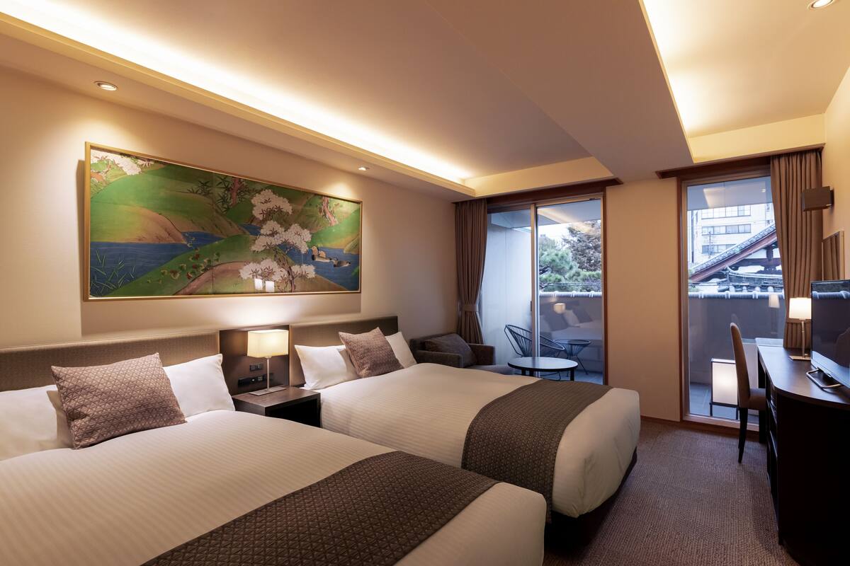 A hotel room with two beds, a desk, a chair, a TV, and a balcony with a view of a building.