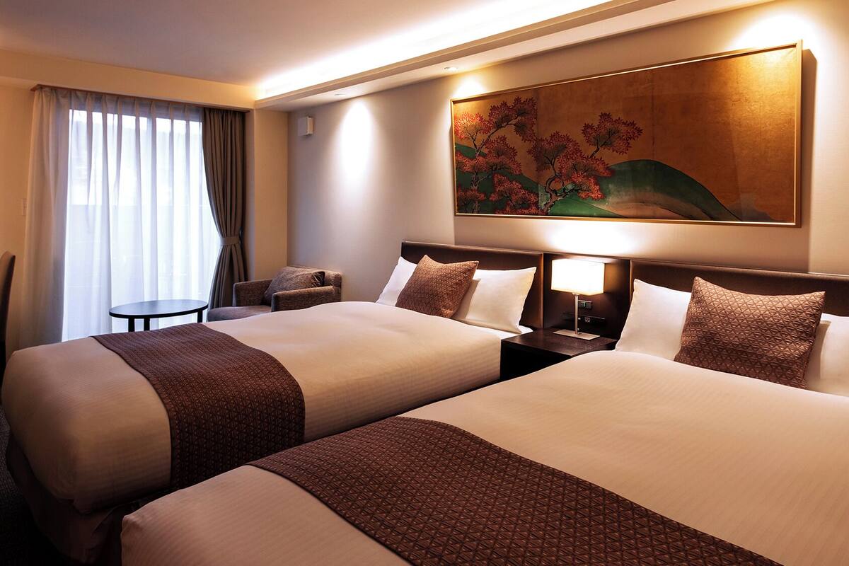 A hotel room with two beds, a desk, a chair, a small table, and a painting on the wall.