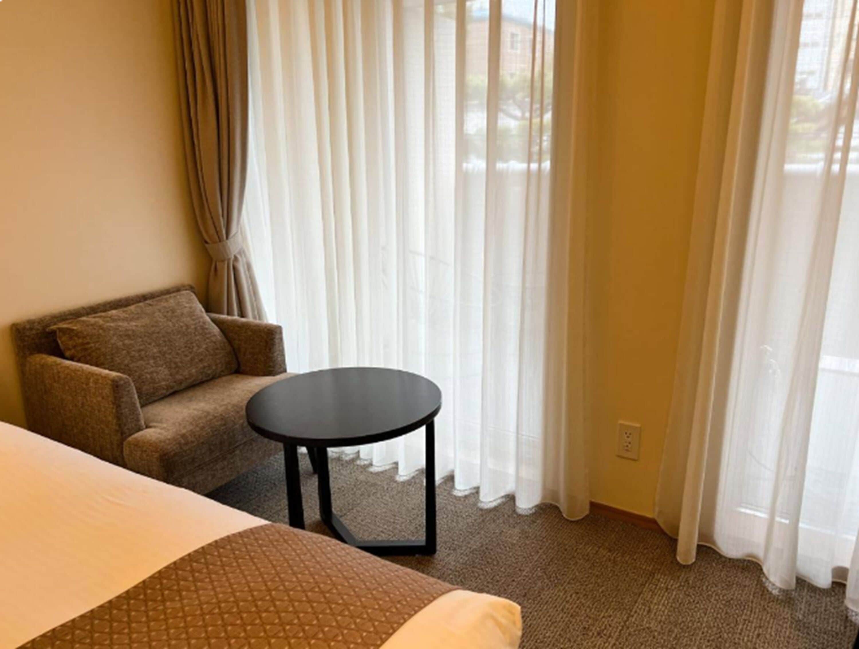 A hotel room with a bed, a sofa, a small round table, and a window with sheer curtains.
