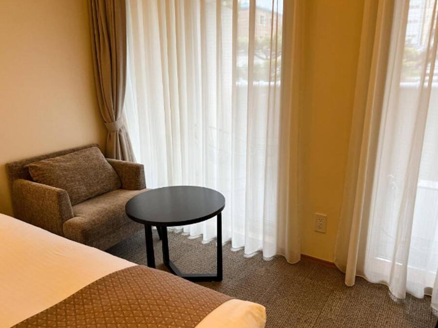 A hotel room with a bed, a sofa, a small round table, and a window with sheer curtains.