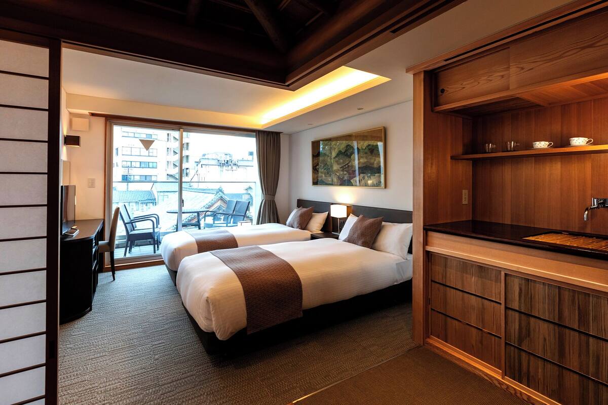 A hotel room with a bed, a desk, a chair, a view of buildings, and a wooden cabinet.