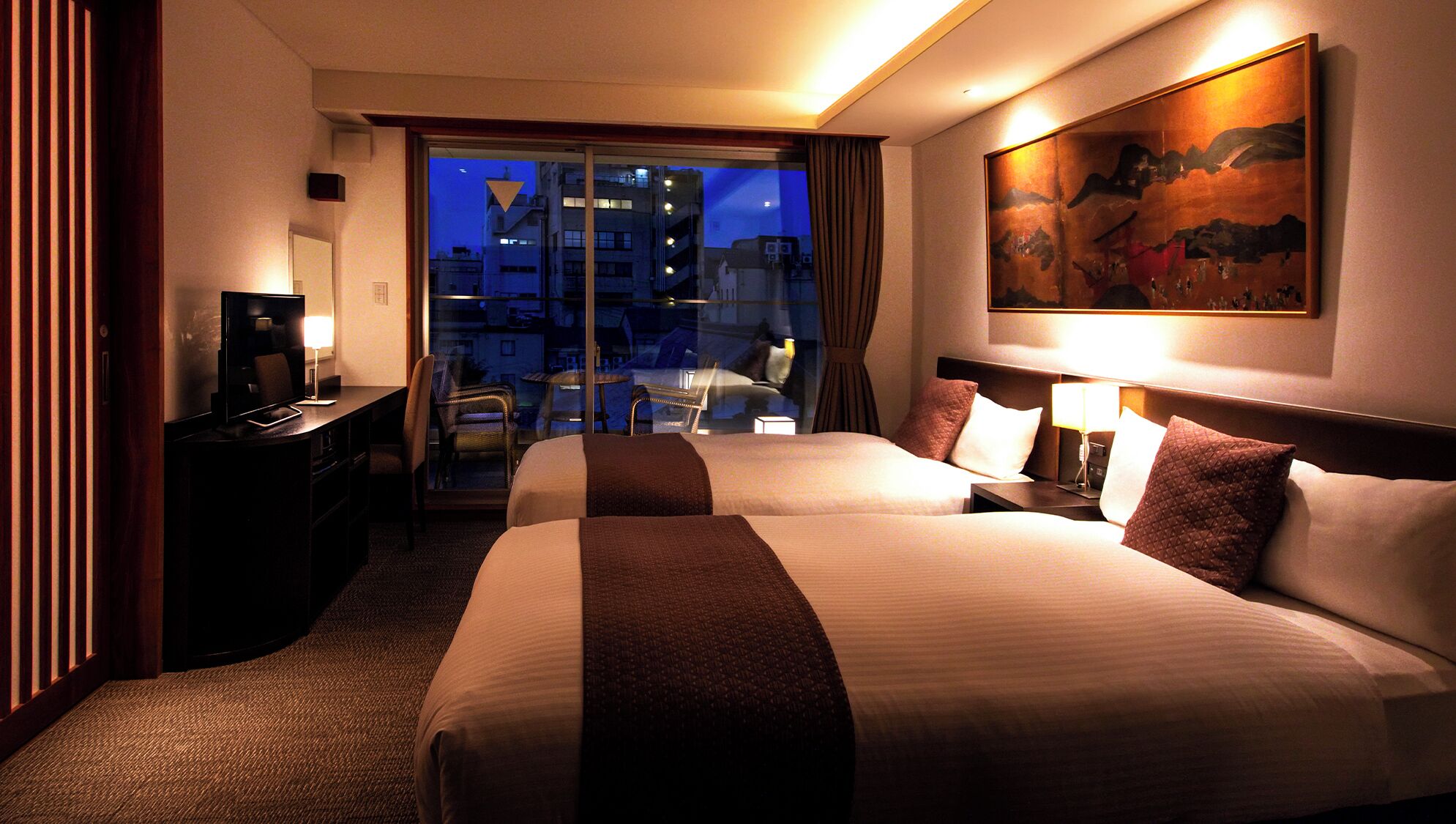 Quarto de hotel com duas camas, uma escrivaninha, uma cadeira, uma televisão e um quadro na parede.
