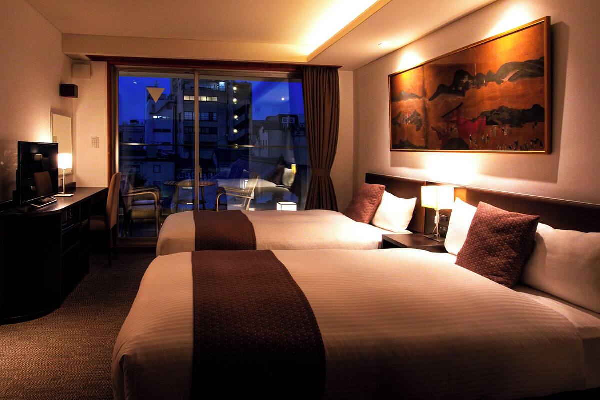 A hotel room with two beds, a desk, a chair, a TV, and a painting on the wall.