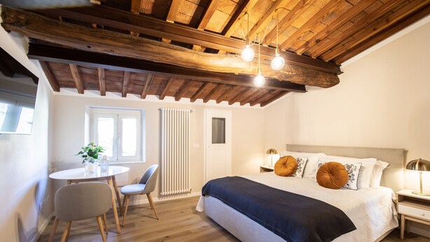 A bedroom with a wooden ceiling, a bed with white bedding and orange pillows, a bedside table with a lamp, and a window with blinds. - Sibilla Luxury Flat in center town (Lucca)
