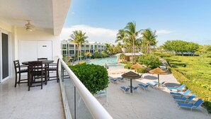 Un balcón con mesa y sillas, con vista a una piscina con sillones y sombrillas, rodeado de palmeras y un edificio con varias ventanas.