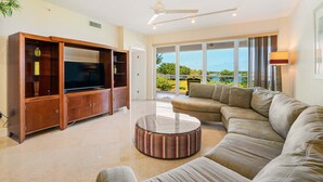 Una sala de estar con un mueble de entretenimiento grande de madera, un sofá color beige, una mesa redonda y un amplio ventanal corredera que da a un balcón con vistas al océano.