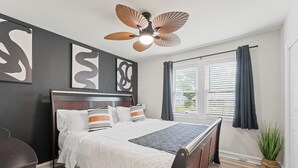 Un dormitorio con cama, cabecera de madera oscura, ventilador de techo, obras de arte, ventana con cortinas y una planta en maceta.