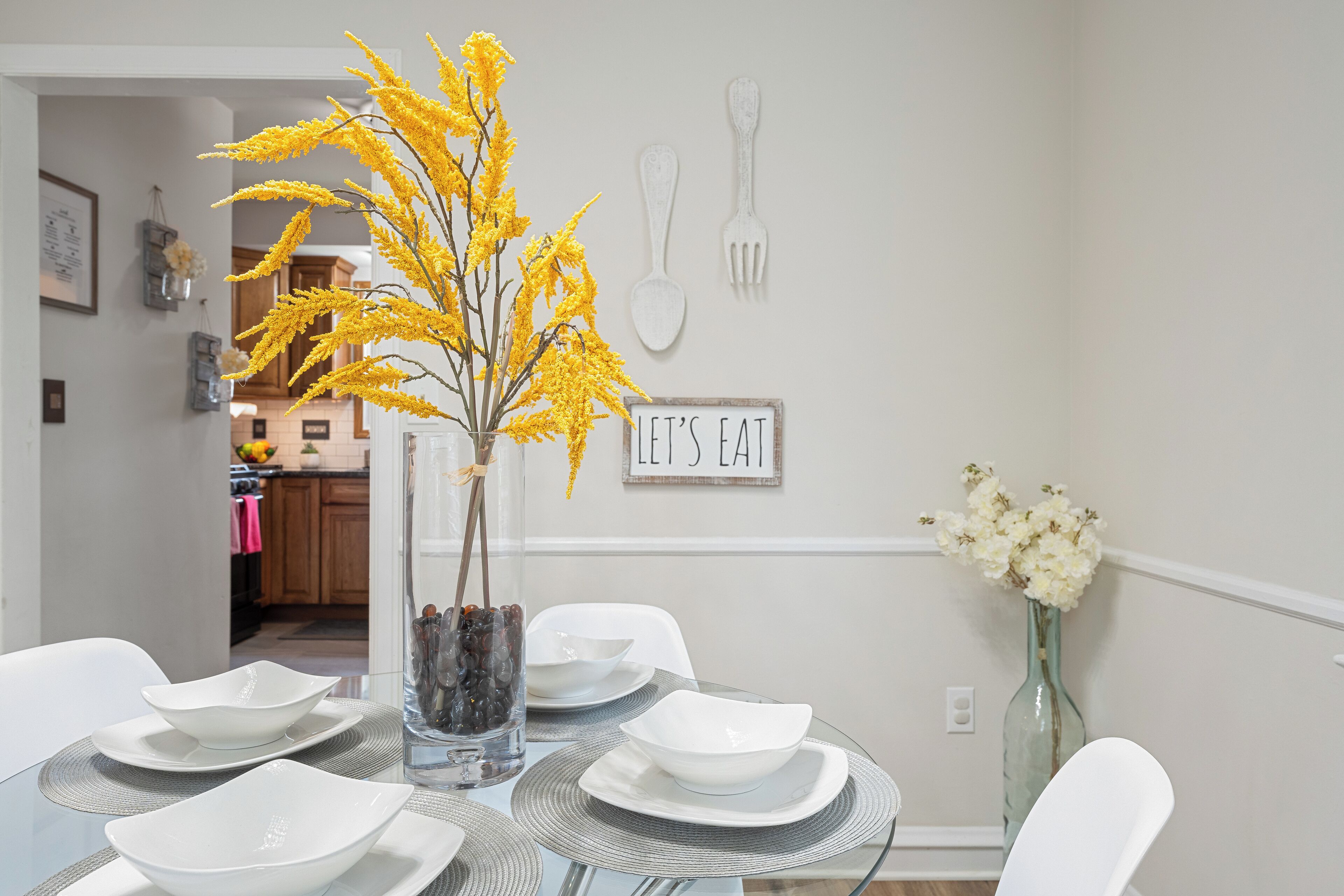 A dining table set with white plates and bowls, a glass vase with yellow flowers, and a 'LET'S EAT' sign on the wall.