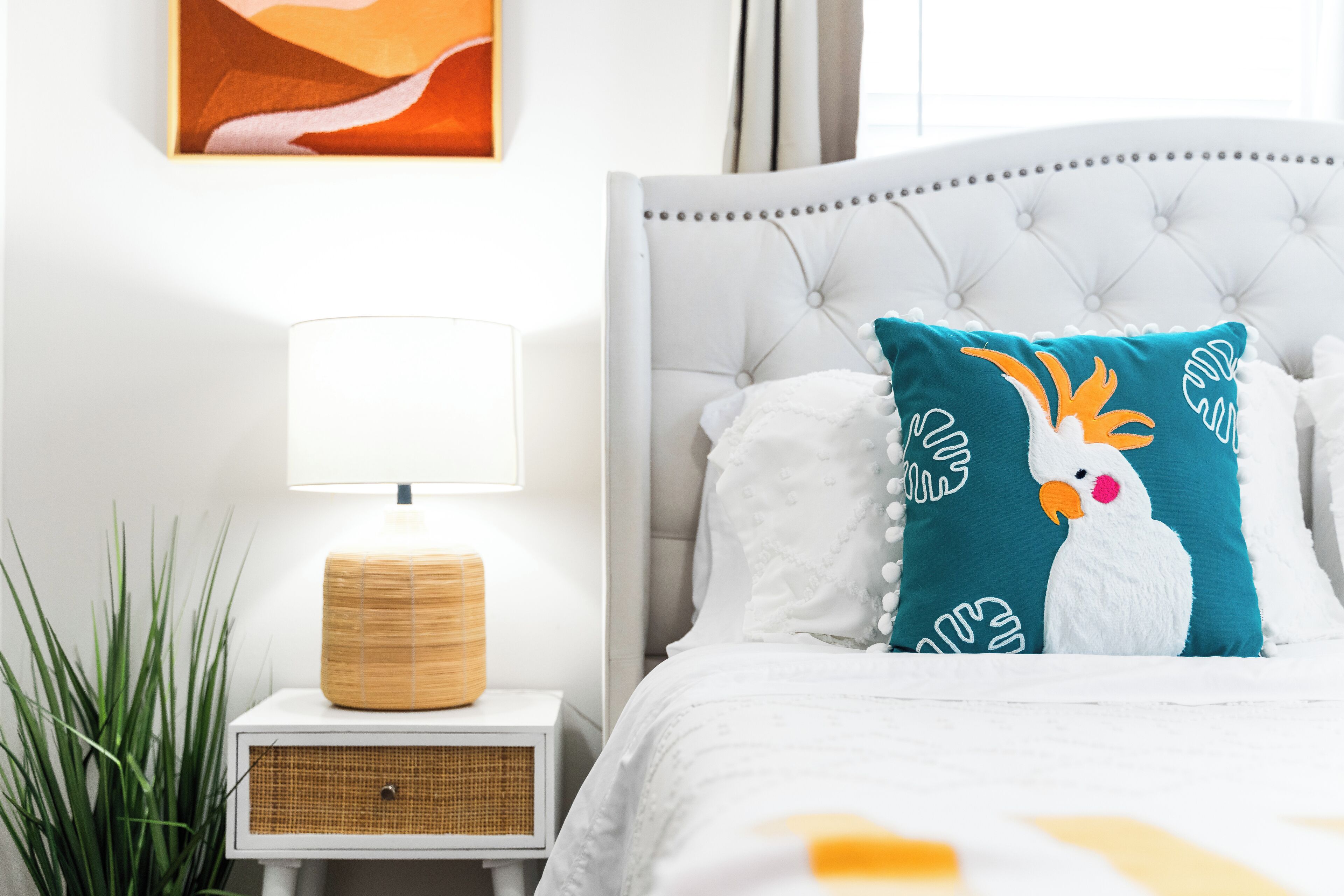 Une chambre à coucher bien rangée, avec un lit blanc, un coussin turquoise orné d’un motif d’oiseau, une lampe de chevet blanche et une table de chevet en osier.