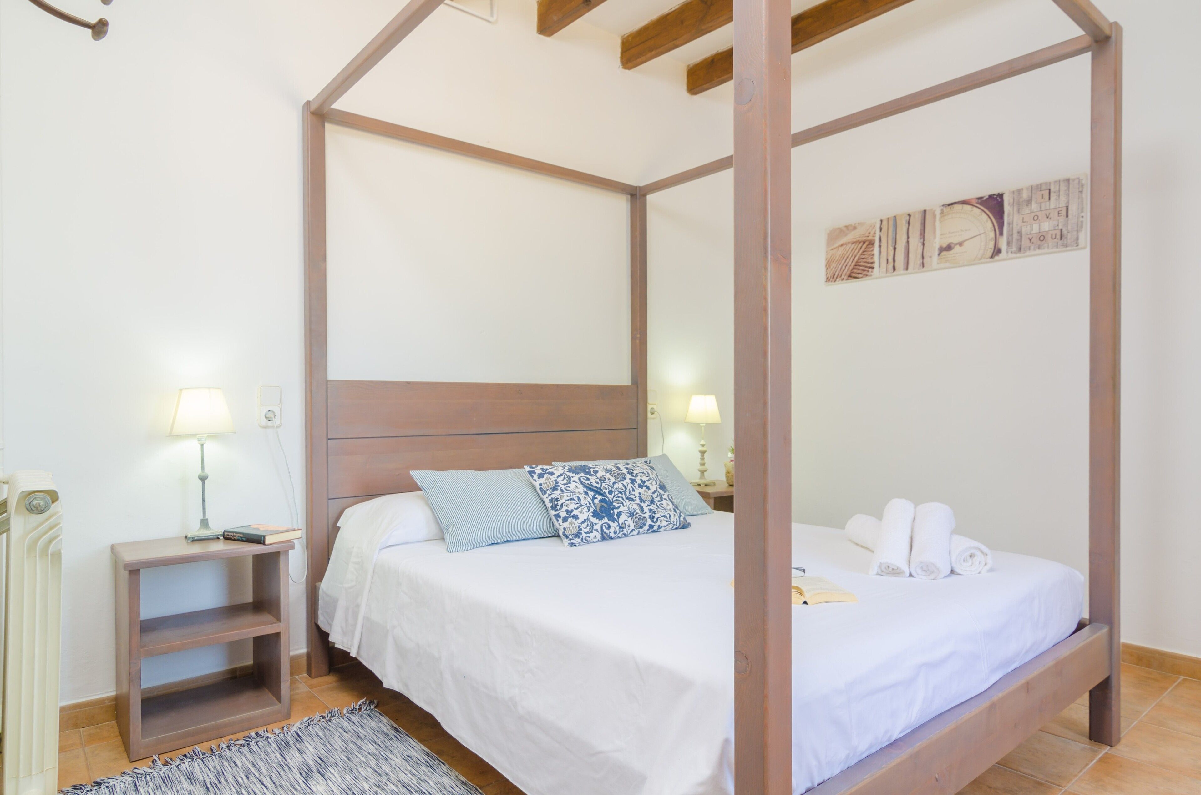 Un lit à baldaquin avec du linge de lit blanc et des coussins bleus, une table de chevet en bois avec une lampe, et un petit tapis sur le sol.
