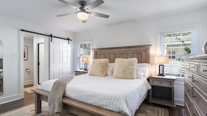 Une chambre à coucher comprenant un lit, des tables de chevet, une commode, une coiffeuse, un ventilateur de plafond et une fenêtre avec des stores.