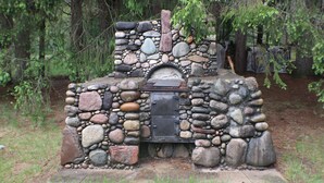 A stone-built outdoor fireplace with a metal door, surrounded by a natural setting with trees and grass. - Cozy Countryside Stone House & Pub with 20 acres of outdoor fun! (Morley)