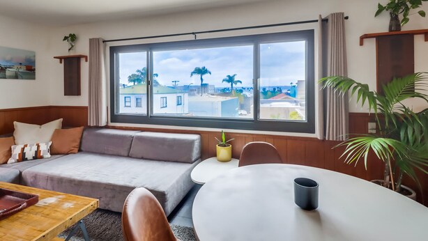 A modern living room with a large window, a sofa, a dining table, and a potted plant.