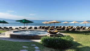 Zona de piscina con sillones de descanso y sombrillas, un jacuzzi y vista al océano.
