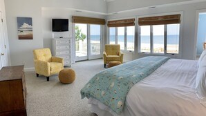 Ein Schlafzimmer mit einem großen Bett, zwei Sesseln, einer Kommode und einem Fernseher. Große Fenster bieten Blick auf den Ozean.
