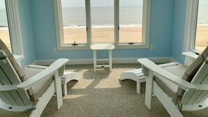Ein Zimmer mit großem Fenster mit Blick auf einen Strand, zwei weiße Stühle und ein kleiner Tisch.