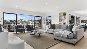 A spacious living room with a grey sofa, two armchairs, a coffee table, and a large window offering a view of the city. - Luxury Mission Bay Oasis with Heated Pool (Auckland)