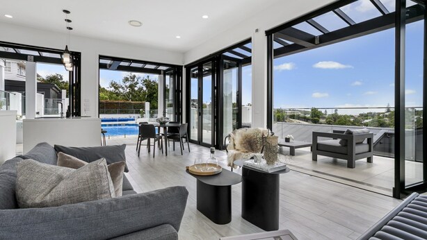 A spacious living area with a modern design, featuring a grey sofa, a dining table with chairs, and a view of a swimming pool through large glass doors. - Luxury Mission Bay Oasis with Heated Pool (Auckland)