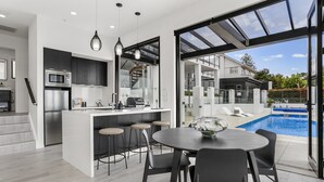 A modern kitchen with a central island, bar stools, and a view of a swimming pool. - Luxury Mission Bay Oasis with Heated Pool (Auckland)