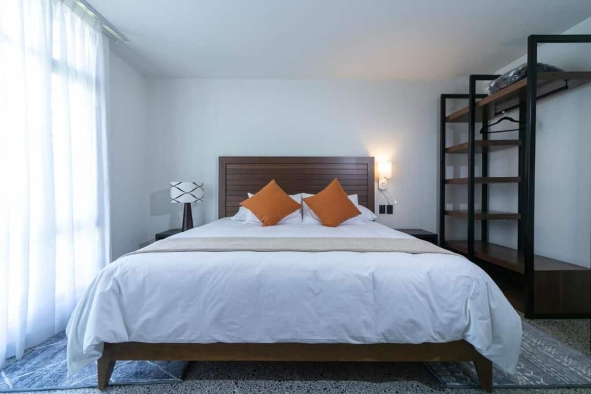 A neatly made bed with white bedding and two orange pillows, a wooden headboard, a bedside table with a lamp, and a built-in shelving unit to the right.