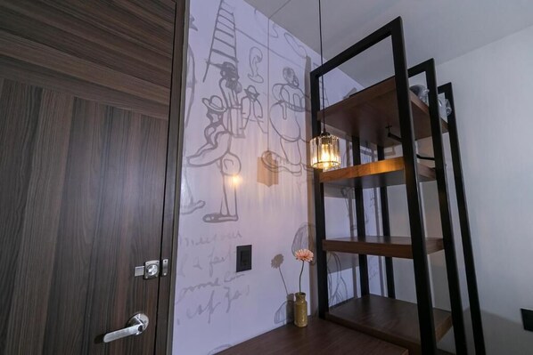 A room with a wooden door, a wall with abstract drawings, a shelf with a vase, and a hanging light fixture.