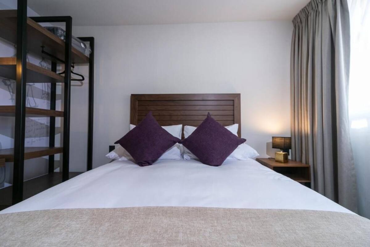 A hotel room with a wooden headboard, two purple pillows, a white bedspread, a nightstand with a lamp, and a wardrobe with a mirror.