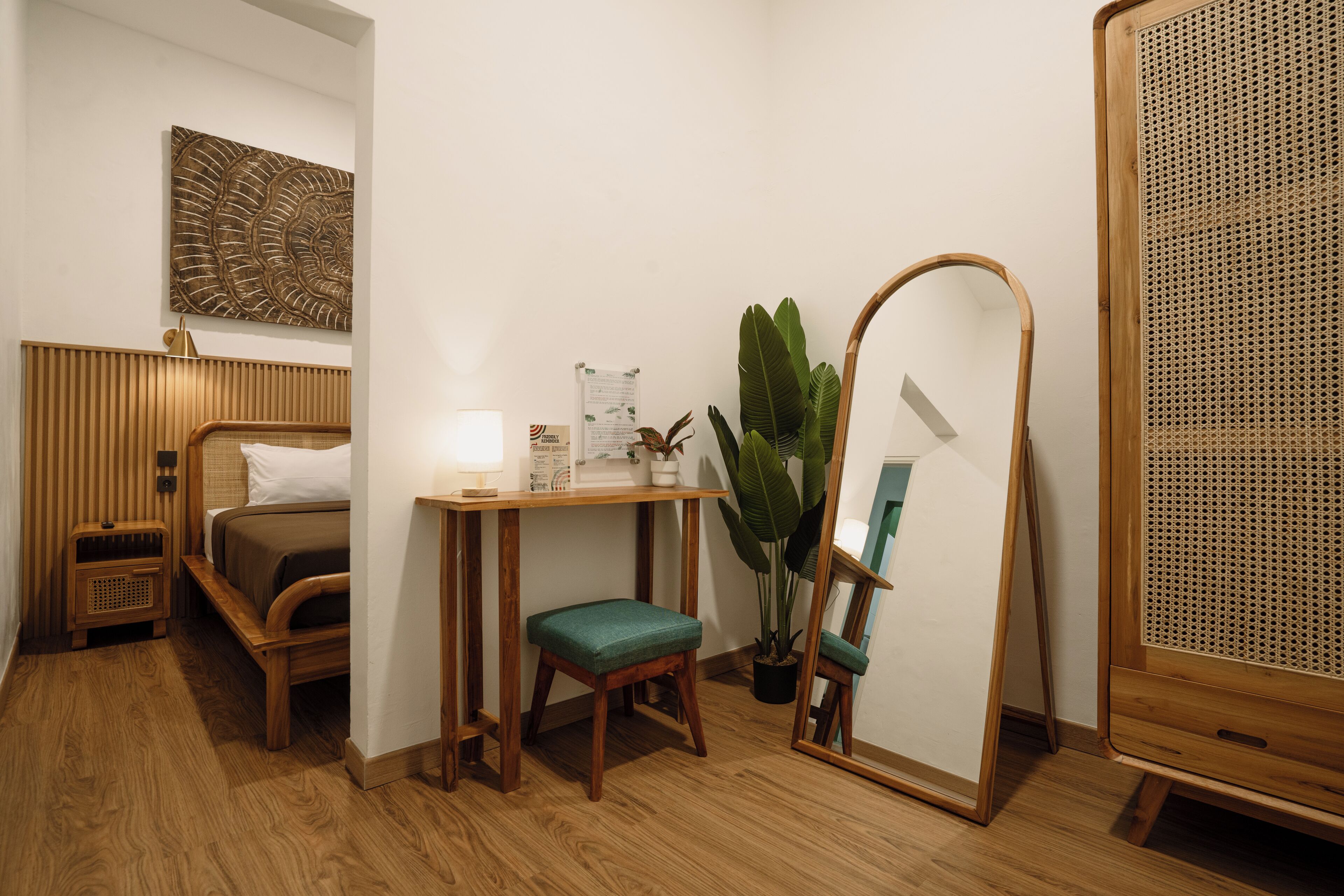 Een compacte hotelkamer met een bed, een klein bureau met een lamp, een spiegel, een potplant en een rieten stoel.